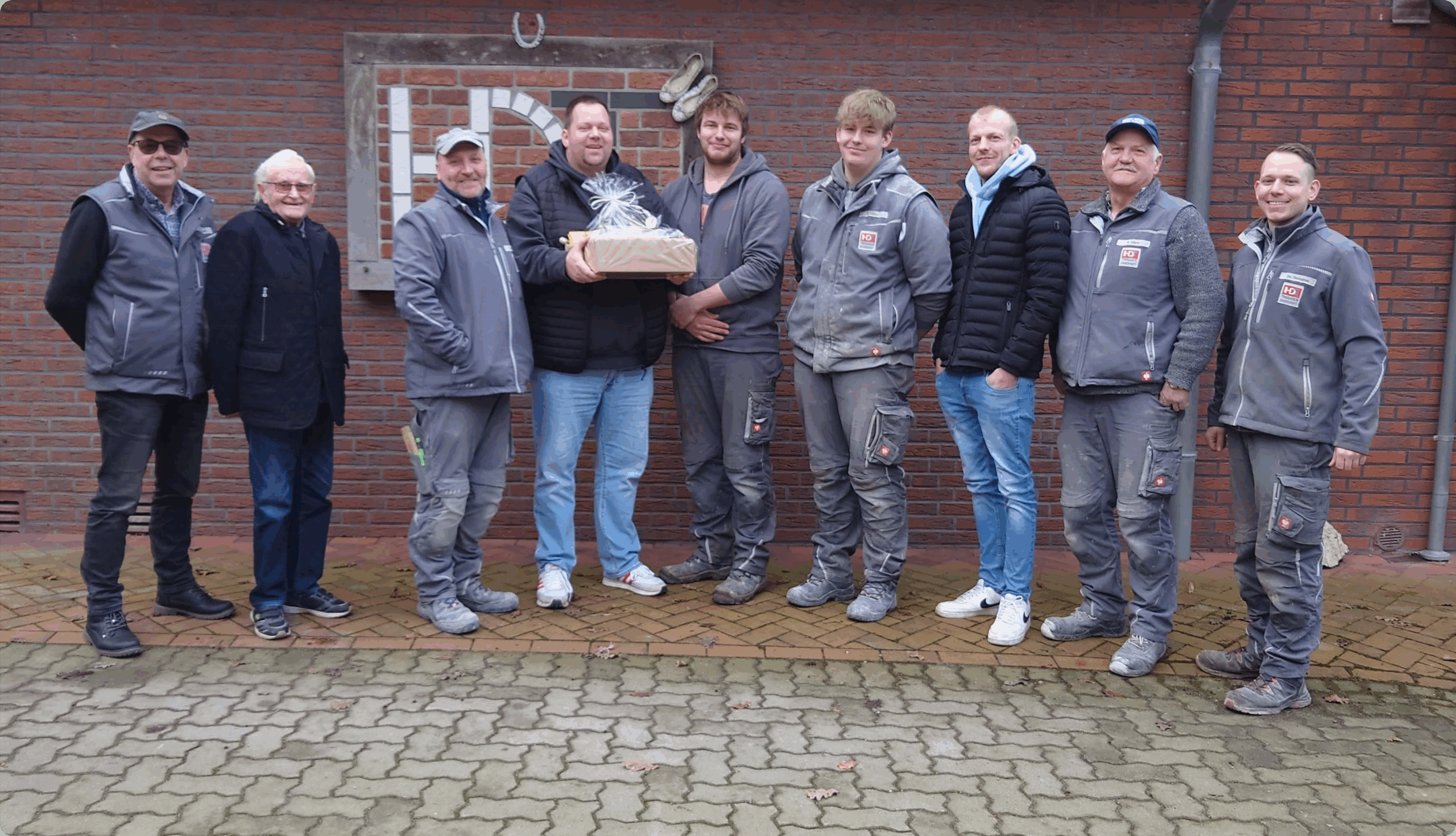 Gruppenfoto eines Handwerkerteams vor einer Klinkerwand mit HDT-Logo und Geschenkkorb.