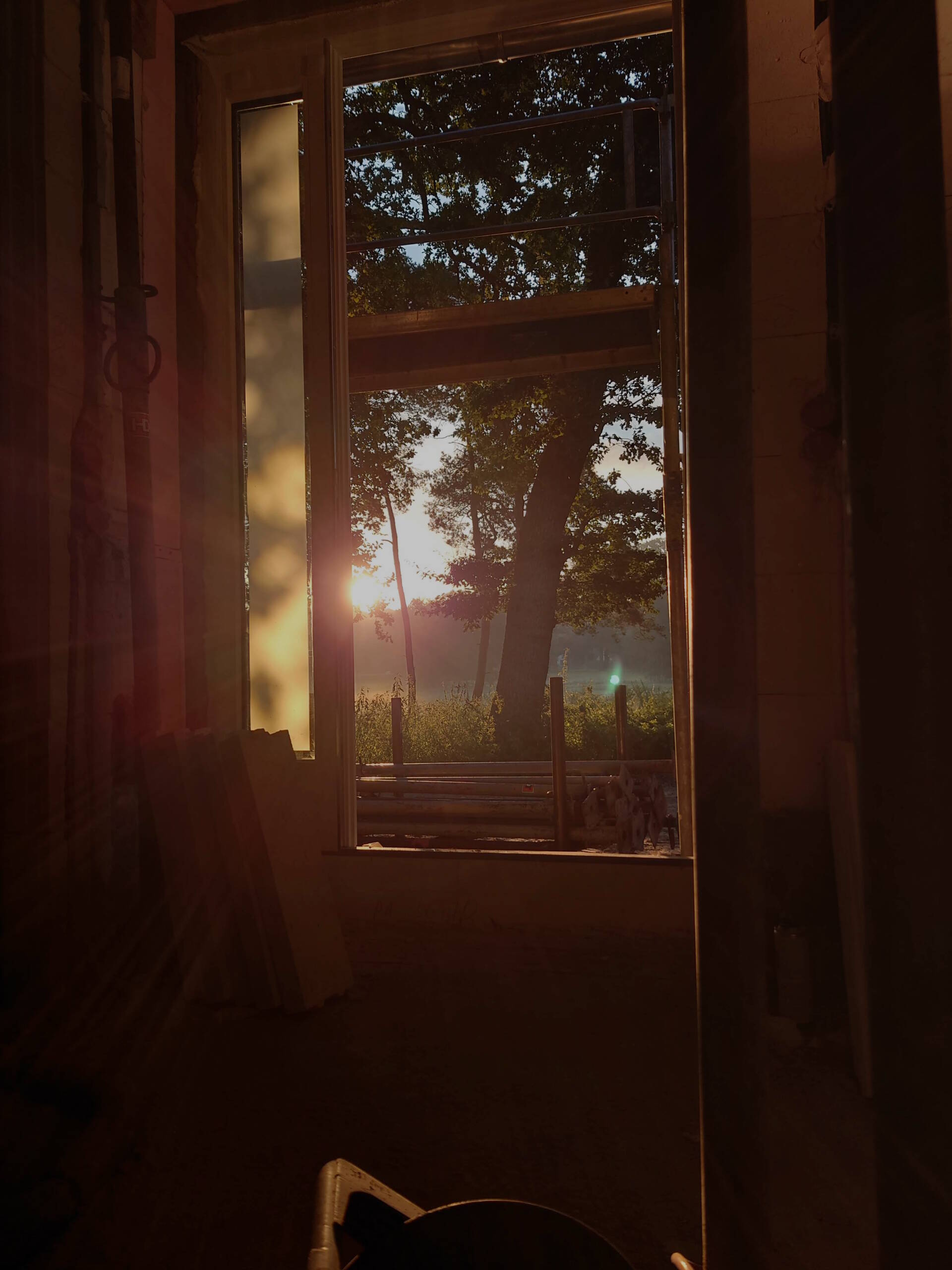 Blick aus einem Rohbaufenster in eine morgendliche Landschaft mit Sonnenaufgang.