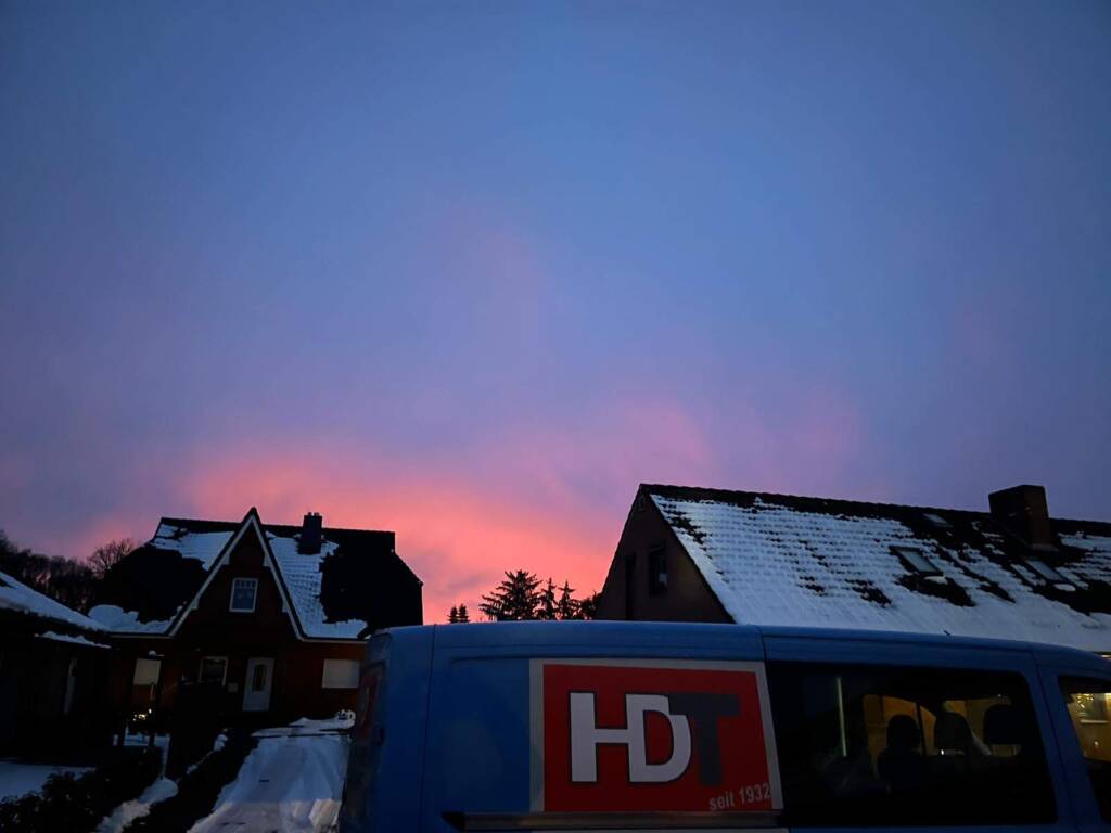 Sonnenuntergang mit pink-blauem Himmel über verschneiten Hausdächern – im Vordergrund ein Fahrzeug mit HDT-Logo.