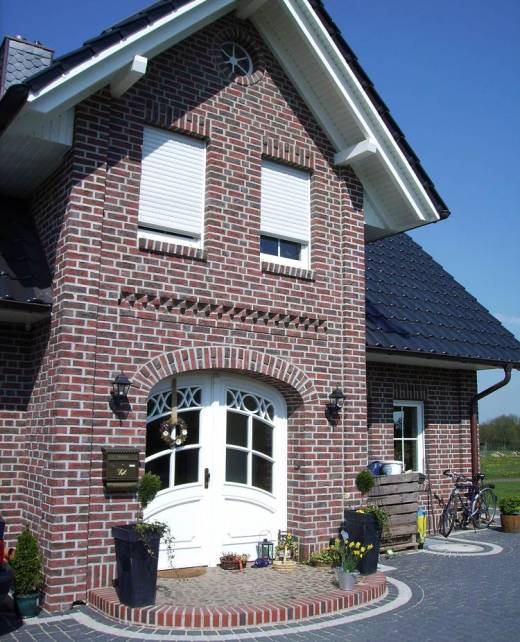 Eingangsbereich eines modernen Einfamilienhauses mit rotem Klinker, Rundbogen über der weißen Haustür und dekorativ bepflanzter Fläche.